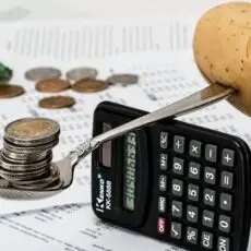 Symbolické znázornění inflace: mince na lžíci opřené o kalkulačku a bramboru, doplněné finančními dokumenty.